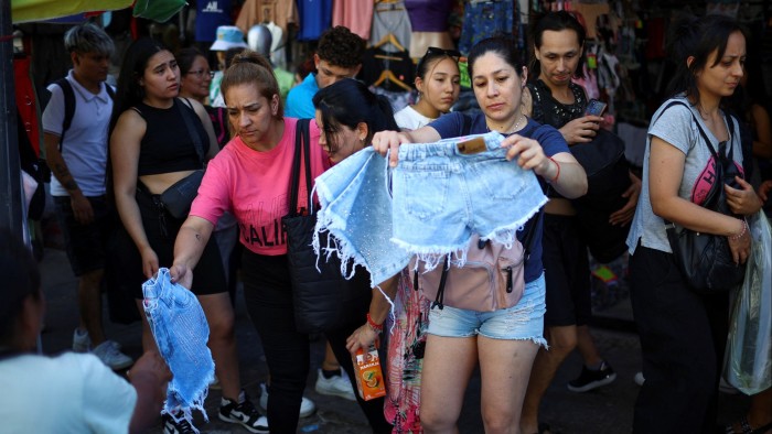 A woman holds up a pair of cut-off denim shorts. Other shoppers are milling around