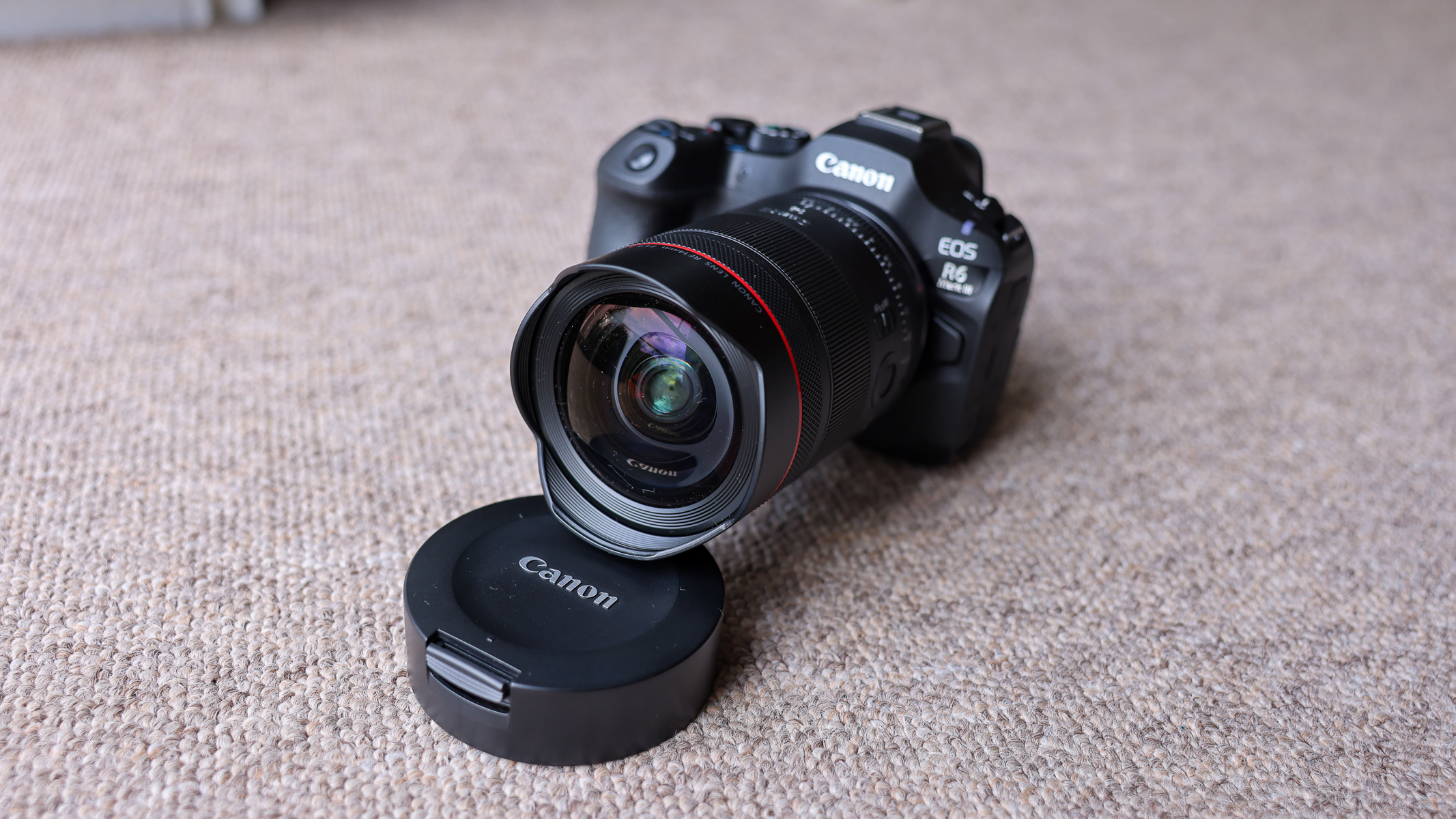 The Canon EOS R6 III with the Canon RF 14mm f/1.4 lens attached and propped up by the lens cover, on a brown carpet.