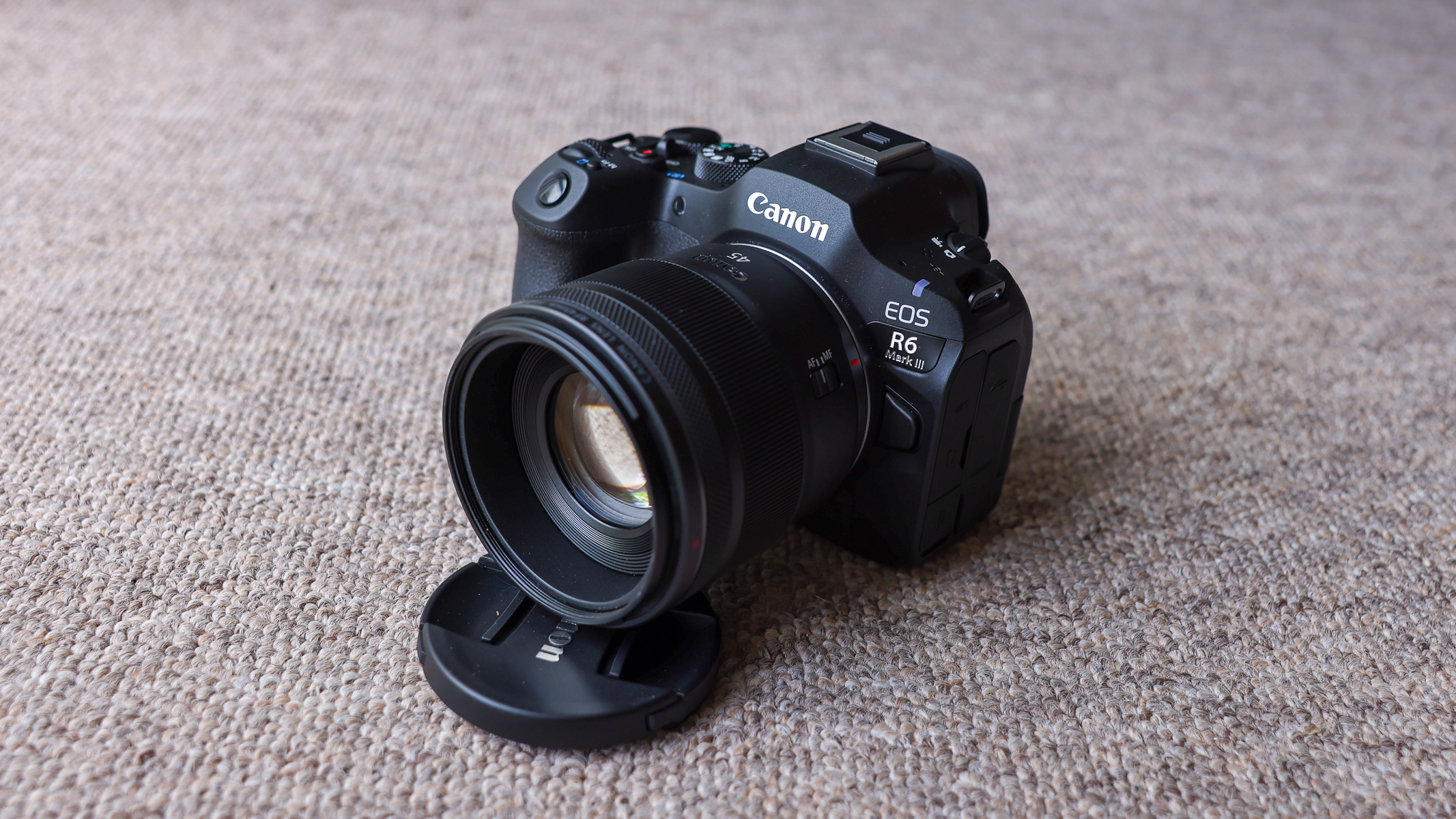 The Canon EOS R6 III with the Canon RF 45mm f/1.2 lens attached and propped up by the lens cover, on a brown carpet.